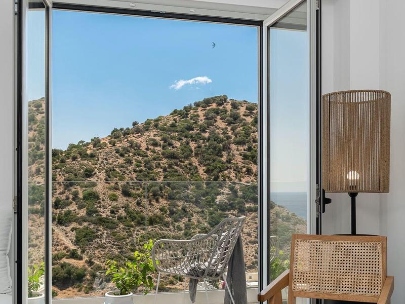 Open raam met uitzicht op heuvels, terras met stoelen en tafel, houten stoel binnen.