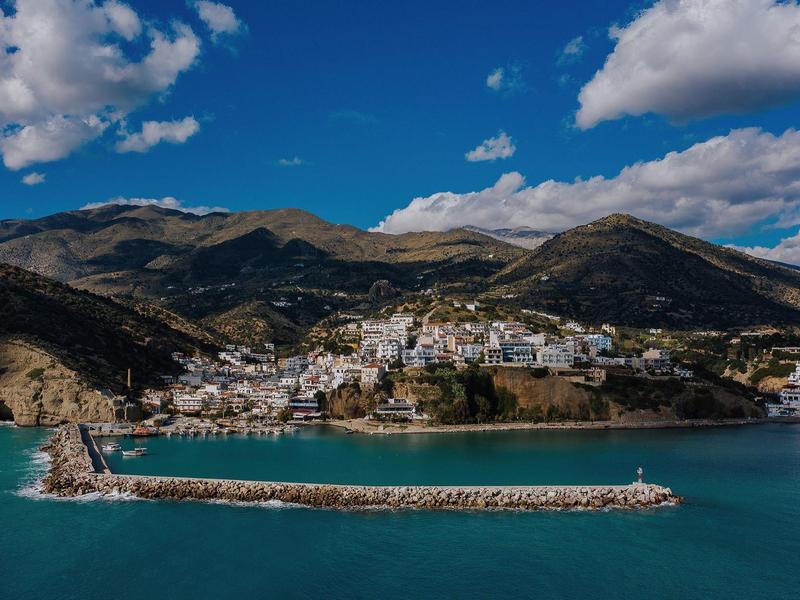 Klein kustdorp met haven, omringd door bergen en blauwe lucht.