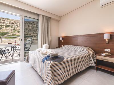 Helles Schlafzimmer mit Doppelbett, braunem Kopfteil, Nachttisch und Zugang zu Balkon mit Tisch und Stühlen.