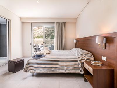 Helles Schlafzimmer mit Doppelbett, Holzkopfteil, Nachttisch, Sitzhocker und Balkon mit Blick auf Felsen.