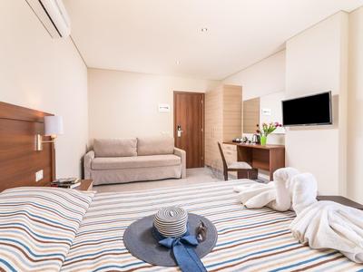 Hell eingerichtetes Hotelzimmer mit Doppelbett, Sofa, Schreibtisch, Wand-TV und modernem Holzmobiliar.