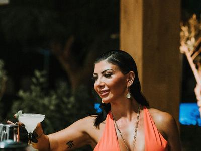 Mujer con top naranja sosteniendo una copa de cóctel en un ambiente exterior acogedor.