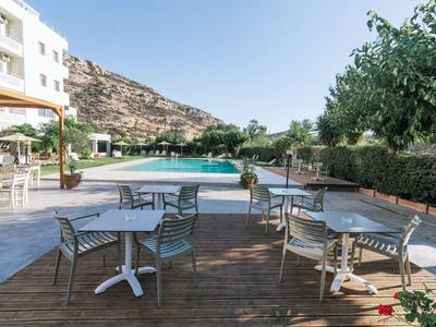 Zona de estar con mesas y sillas en una terraza de madera junto a una piscina con montañas al fondo.