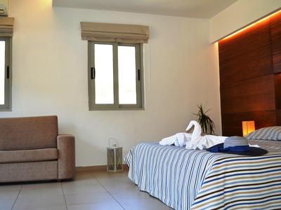 Helles Schlafzimmer mit Doppelbett, gestreifter Bettwäsche, Sofa und zwei Fenstern mit Jalousien.