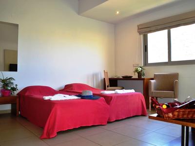 Helles Schlafzimmer mit zwei roten Betten, kleinem Tisch, Stuhl und Fenster mit Rolladen.