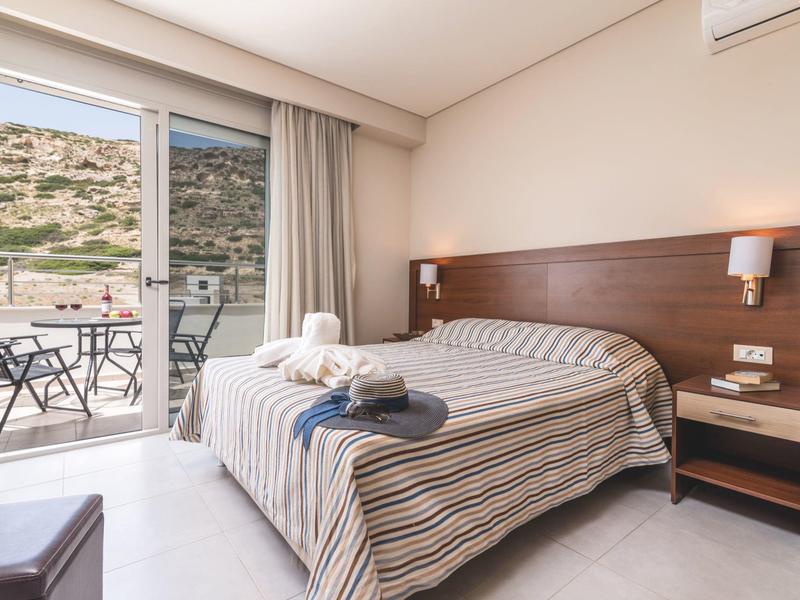 Helles Schlafzimmer mit Doppelbett, braunem Kopfteil, Nachttisch und Zugang zu Balkon mit Tisch und Stühlen.