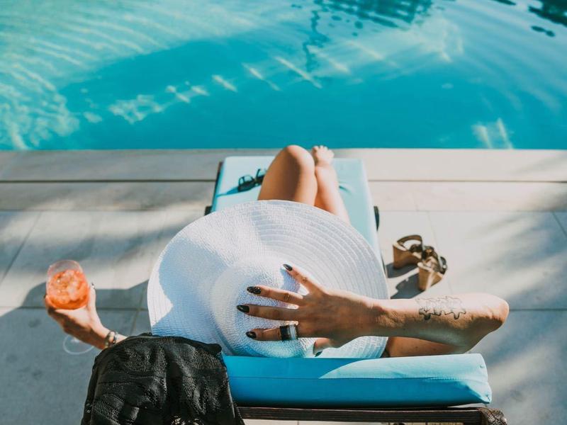 Person mit weißem Kleid liegt entspannt auf blauem Liegestuhl am Pool, hält Drink in Hand.