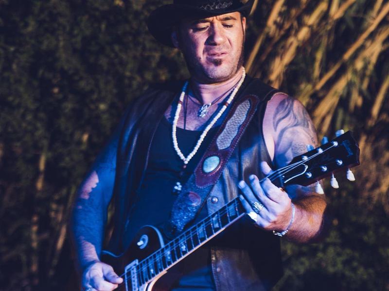 Mann spielt Gitarre, trägt Weste und Hut, dunkler Hintergrund mit Bäumen und Lichtakzenten.