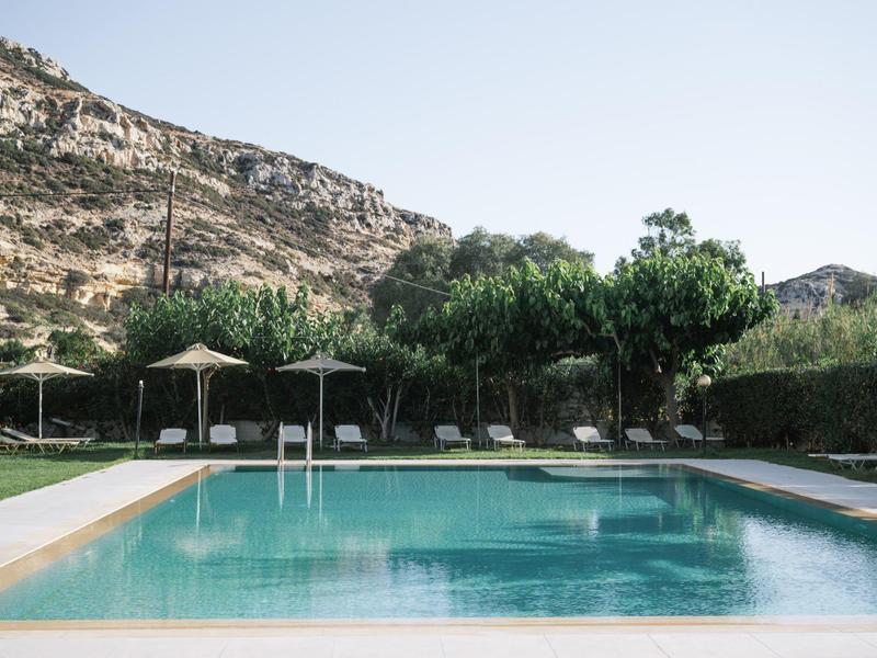 Ein ruhiger Pool mit Liegen, Sonnenschirmen und Bäumen vor felsigem, sonnenbeschienenem Hügel.