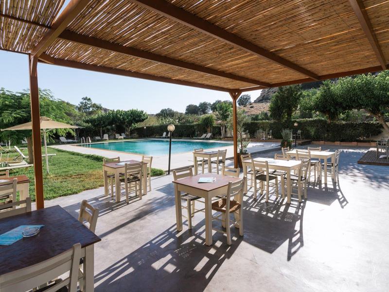 Terrasse mit Holzdach, mehreren Tischen und Stühlen vor einem Pool in grüner Umgebung.