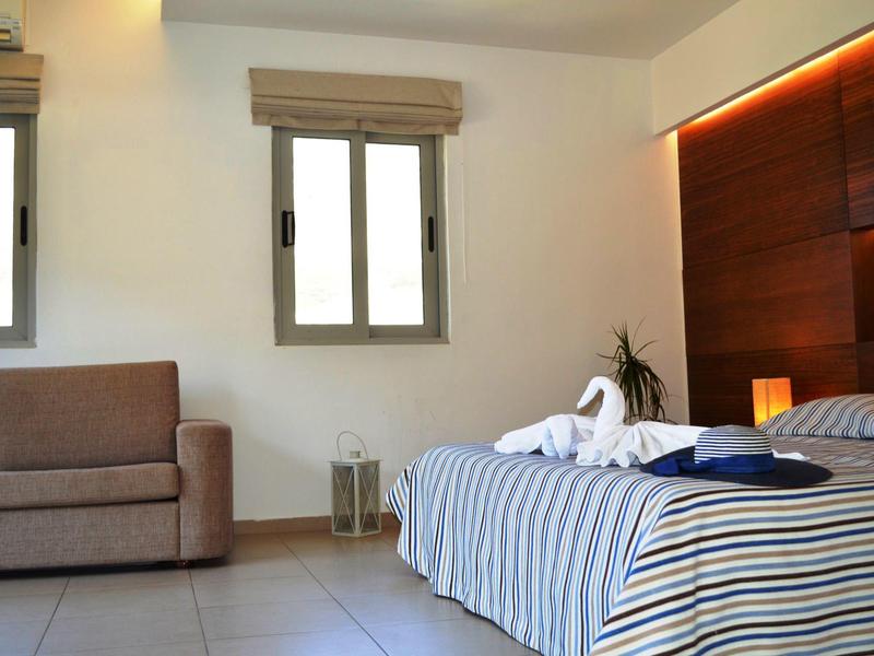 Helles Schlafzimmer mit Doppelbett, gestreifter Bettwäsche, Sofa und zwei Fenstern mit Jalousien.