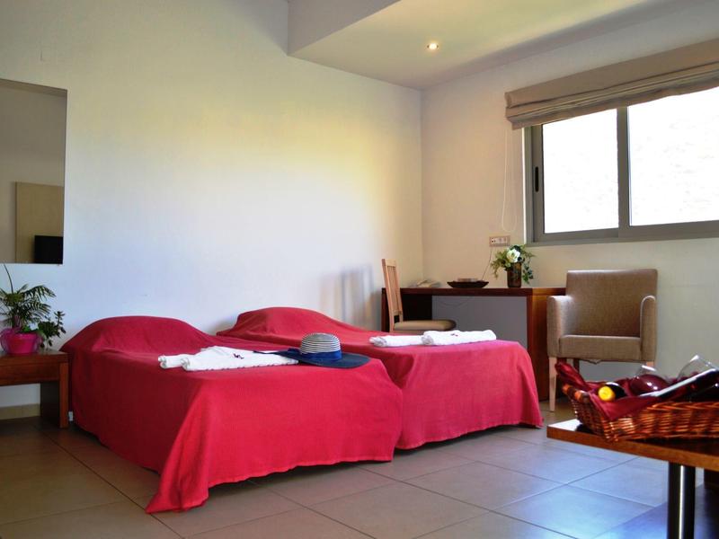 Helles Schlafzimmer mit zwei roten Betten, kleinem Tisch, Stuhl und Fenster mit Rolladen.