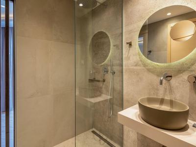 Nowoczesna łazienka z okrągłym lustrem, umywalką i prysznicem typu walk-in w neutralnych kolorach.