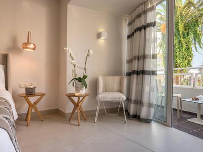 Habitación de hotel luminosa con sillas cómodas, mesas de madera, cortinas y balcón con vista a palmeras.