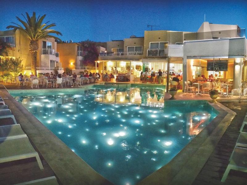 Una piscina iluminada en un hotel por la noche con huéspedes y palmeras de fondo.