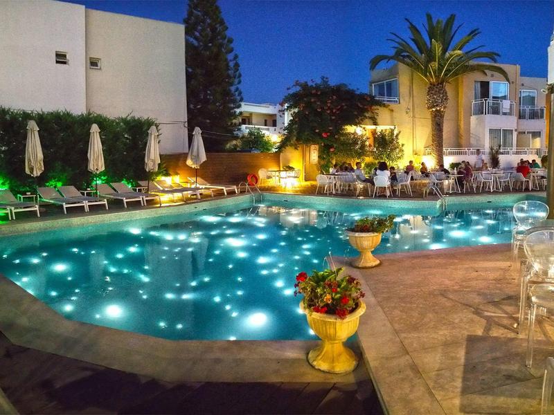 Vista nocturna de una piscina iluminada en un hotel con tumbonas y palmeras al fondo.