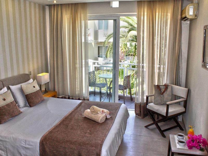 Habitación de hotel luminosa con cama doble, balcón y vista a un jardín con piscina.