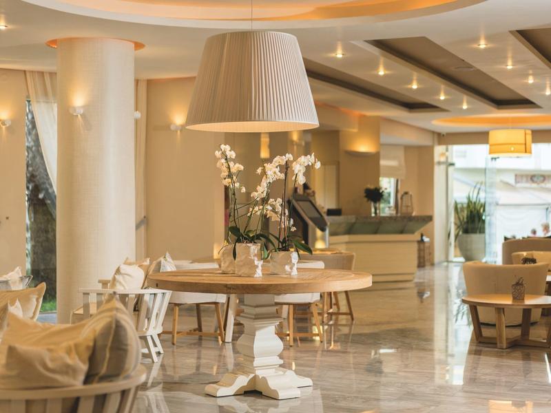 Área de salón elegante en hotel con mesas redondas de madera, sillones cómodos y luz cálida.