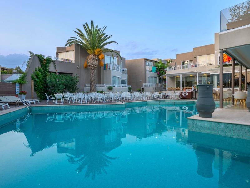 Piscina exterior moderna con tumbonas y palmeras en un hotel bajo cielo claro.