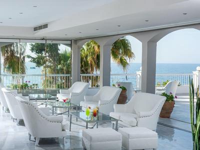 Otwarta przestrzeń wypoczynkowa z białymi fotelami i widokiem na morze na tarasie hotelowym z palmami.