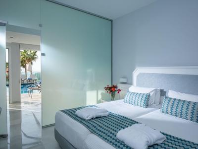 Jasny pokój hotelowy z łóżkiem podwójnym, pasiastą pościelą i widokiem na basen przez szklane przesuwne drzwi.