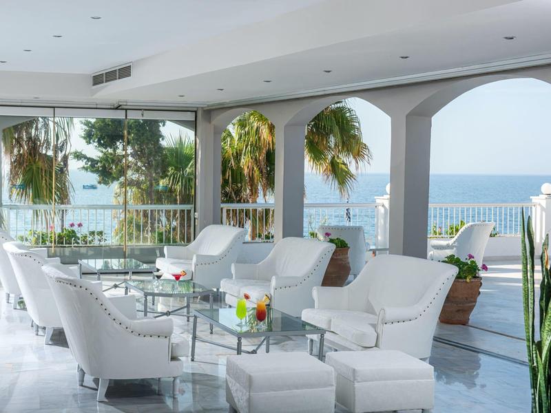 Otwarta przestrzeń wypoczynkowa z białymi fotelami i widokiem na morze na tarasie hotelowym z palmami.