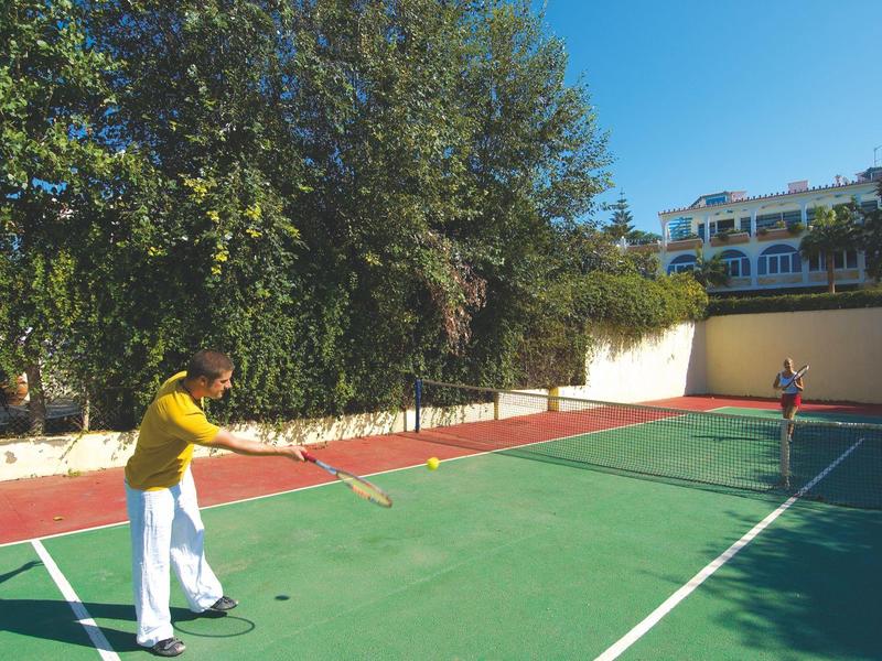 Dwie osoby grają w tenisa na słonecznym korcie przed budynkiem mieszkalnym.