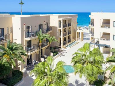 Kompleks hotelowy z nowoczesną architekturą, palmami, basenem i widokiem na morze w tle.