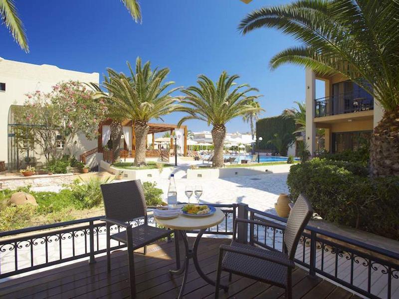 Terrasse mit Stühlen und Tisch, Palmen, blauer Himmel und helle Gebäude im Hintergrund.