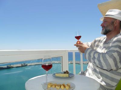 Hombre con sombrero blanco y suéter a rayas disfruta vino en balcón con vista al mar.