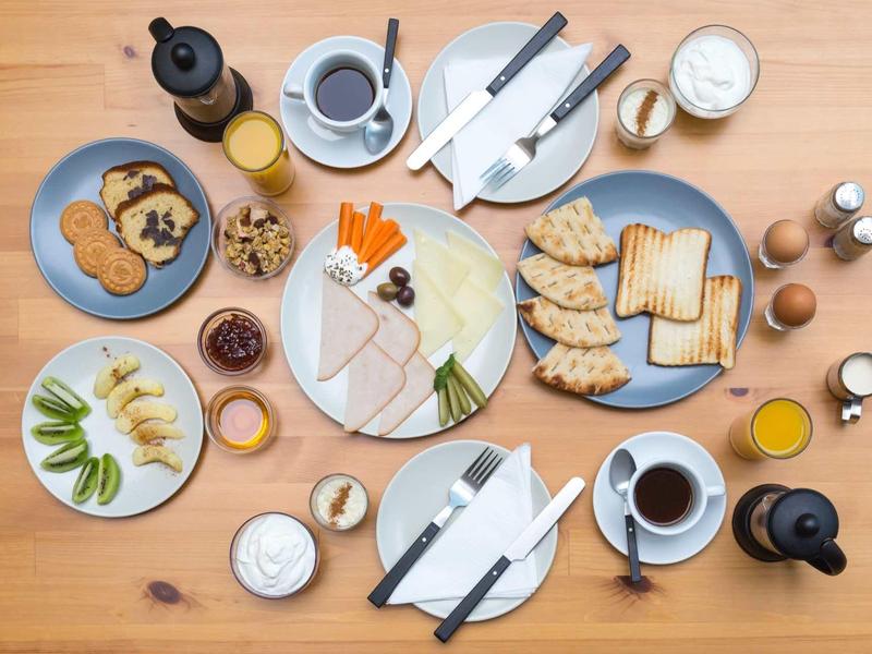 Colazione varia con pasticcini, affettati, verdure, caffè e succo su un tavolo in legno.