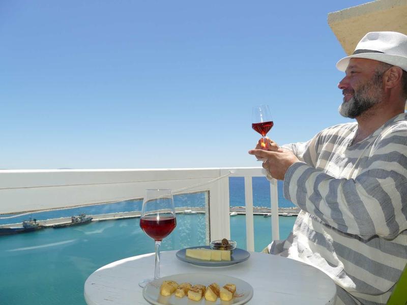 Un uomo si rilassa con un bicchiere di vino rosso su un balcone con vista sul mare e un piatto di formaggi.