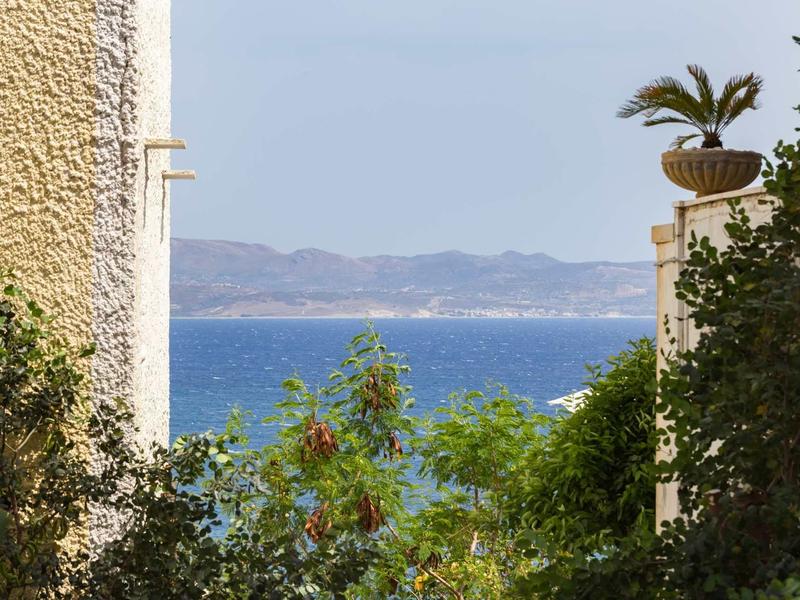 Vista sul mare tra due edifici con piante e montagne sullo sfondo.