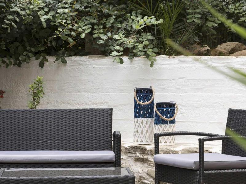 Area esterna moderna con mobili in rattan e cuscini bianchi davanti a un muro bianco con piante verdi.