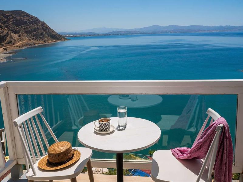 Balcone con tavolo e sedie bianchi con vista sul mare blu e la costa.