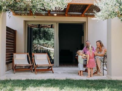 Due donne con due bambini davanti a una terrazza con sedie a sdraio e rami di ulivo.