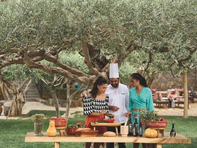 Uno chef e due donne preparano cibo insieme all'aperto sotto gli ulivi.