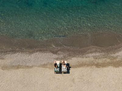 Zwei Personen entspannen auf Liegen am Strand mit klarem, blau-grünem Meer im Hintergrund.