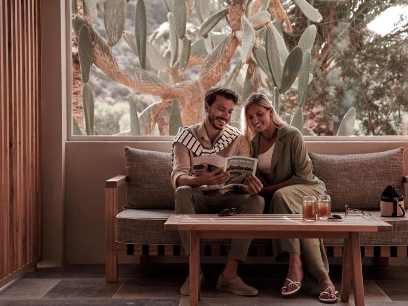 Coppia sorridente seduta su un divano moderno in legno, che legge un libro in un soggiorno luminoso.