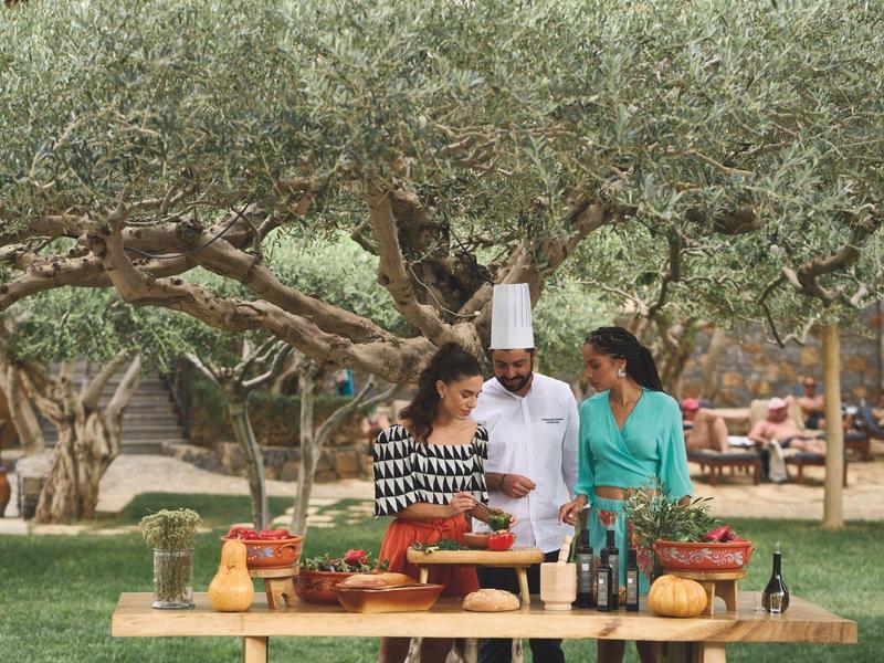 Uno chef e due donne preparano cibo insieme all'aperto sotto gli ulivi.