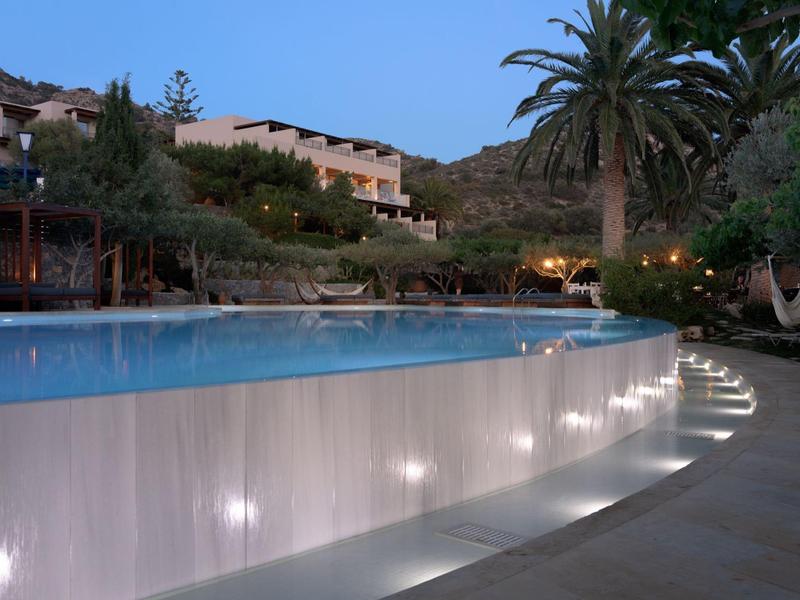 Piscina illuminata di sera con palme e edifici sullo sfondo.