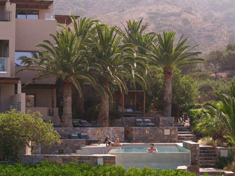 Piscina all'aperto circondata da palme con edificio dell'hotel sullo sfondo, cielo sereno.