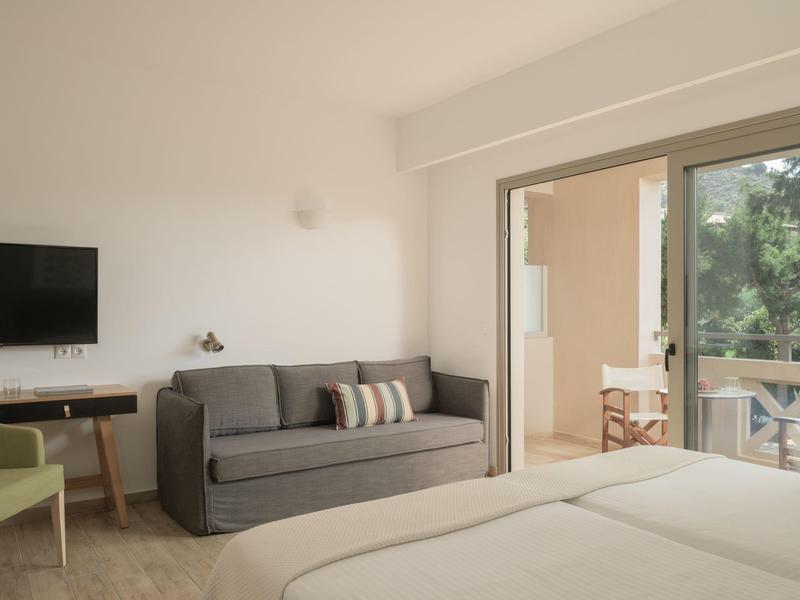 Camera d'hotel luminosa con letto matrimoniale, divano grigio, poltrona verde e balcone aperto.