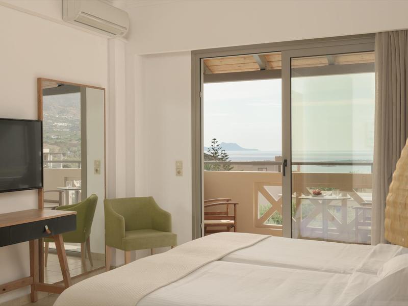 Camera d'hotel luminosa con letto, poltrona verde e terrazza vista mare.