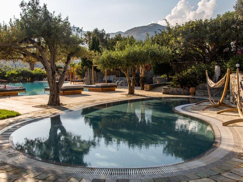 Piscina esterna circondata da alberi e montagne sullo sfondo, con cielo limpido.