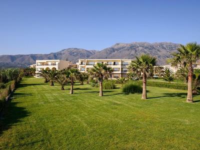 Großes grünes Gartengelände mit Palmen vor modernen Gebäuden und Bergen im Hintergrund.