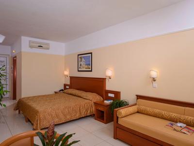 Pokój hotelowy z łóżkiem dwuosobowym, sofą, nocnymi stolikami i roślinami w ciepłych beżowych tonach.