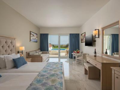 Hell eingerichtetes Hotelzimmer mit zwei Betten, Schreibtisch, Fernseher und Balkon mit Meerblick.