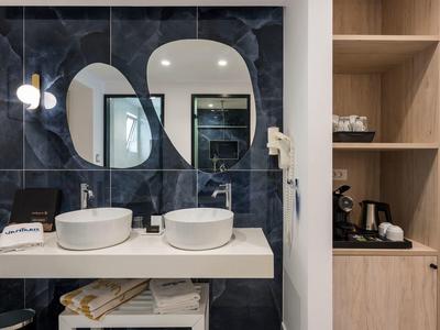 Badezimmer mit zwei weißen Waschbecken, unregelmäßigen Spiegeln und dunklen Wandfliesen.