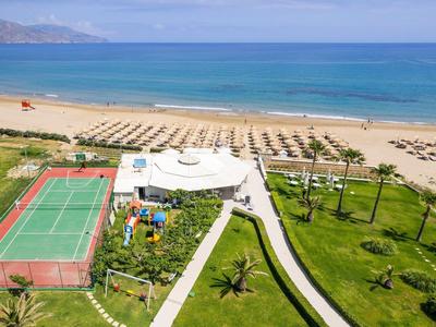 Blick auf Tennisplatz, Poolbereich, Palmen und langen Sandstrand am blauen Meerufer.